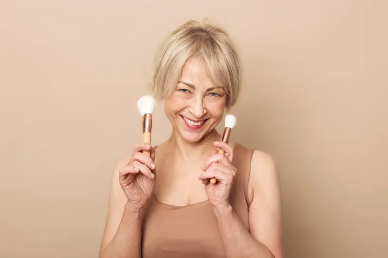 Smiling middle aged woman holding two makeup brushes near face on beige background. Concept of beauty routine, cosmetics, skincare, self care, and natural mature female confidence.