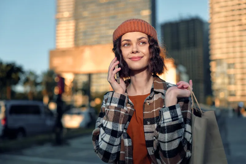 Woman shopping with phone in city, beanie and smile during golden hour, candid lifestyle scene of authenticity and mindful living, emotional storytelling, urban fall fashion.