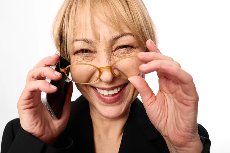 Senior woman with smile adjusts glasses while holding smartphone to ear, phone call communication, friendly portrait, closeup indoor on white background, relaxed hands gesture