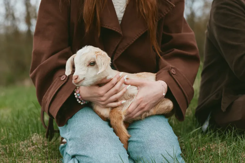 Young woman in brown coat gently holds a small baby goat while sitting on grass outdoors during a casual lifestyle moment in spring season.