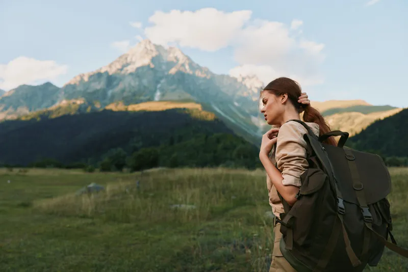 Woman hiker with backpack explores a mountain meadow, embracing outdoor lifestyle, serene scenery, bright sky, distant peaks, casual travel gear, solo adventure