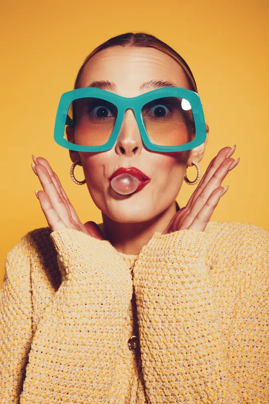 Young woman with oversized turquoise glasses makes a playful face with puckered lips and raised hands. She wears a yellow sweater against a bright yellow background. Fun and quirky portrait.