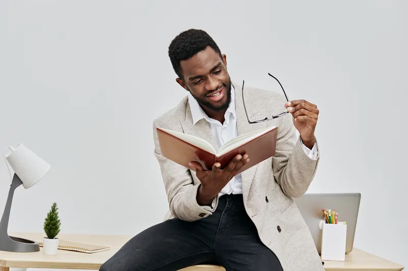 Smiling young man with dark skin and short curly hair, dressed in a beige blazer and white shirt, sitting on a desk holding an open book and glasses, in a modern office environment. People lifestyle c