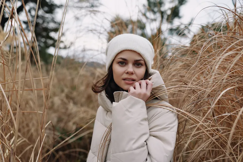 Portrait of a woman in a warm beige coat and white beanie standing in tall grasses, outdoor scene with soft natural light and calm candid mood.