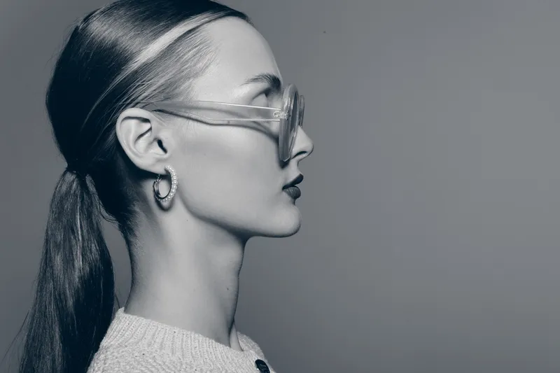 Profile of a young white woman with tied hair and transparent glasses in a grayscale studio portrait. Concept of fashion, accessories and minimalism.