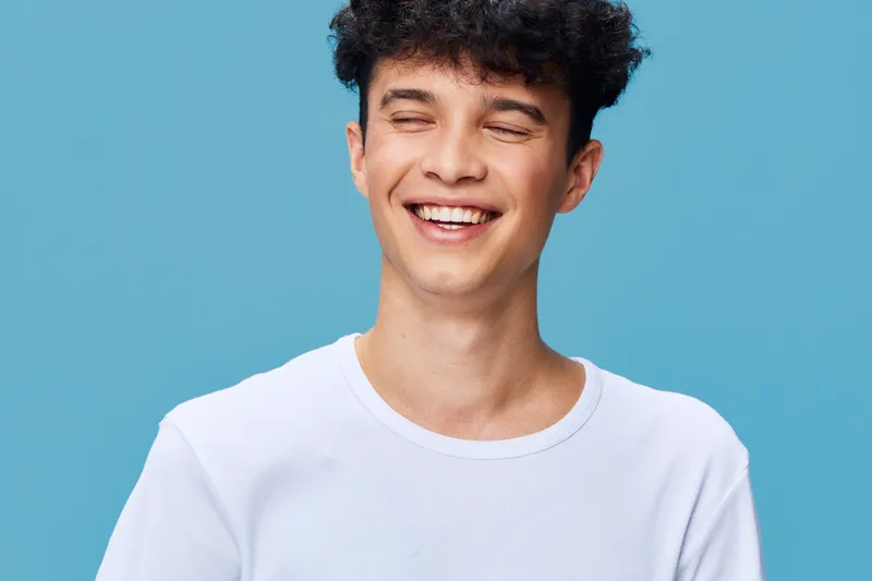 smiling man wearing white t-shirt with curly hair against blue background expressing happiness and joy in a bright studio setting