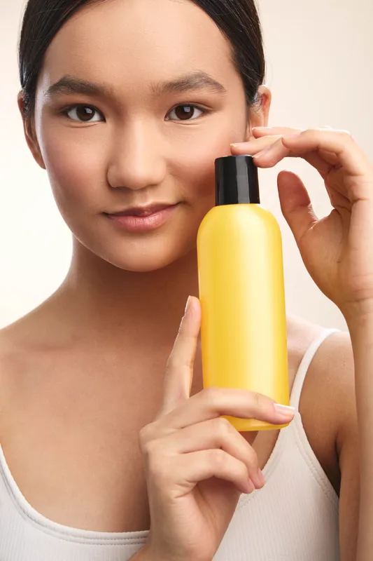 Asian woman holds a yellow cosmetic bottle in a closeup portrait. The neutral backdrop and soft lighting create a clean, fresh beauty moment with a gentle, natural smile.