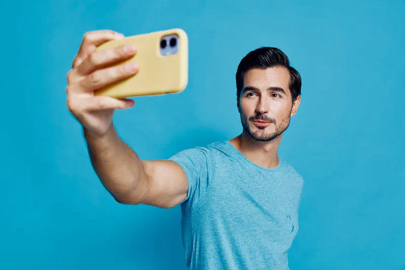 Young man taking selfie with mobile phone, wearing casual light blue t-shirt, posing against solid bright blue background. Modern lifestyle concept.