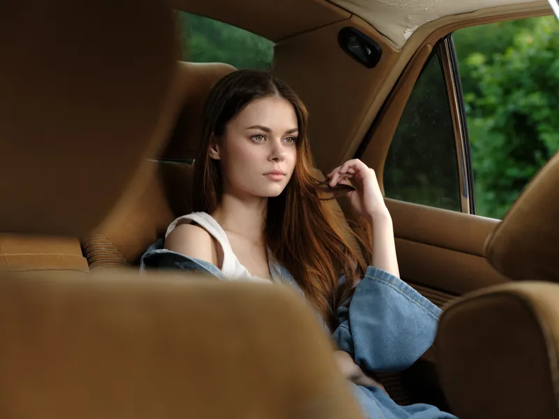 Thoughtful young woman relaxing in car interior, wearing denim jacket, capturing peaceful moment in nature with soft lighting and warm color tones.