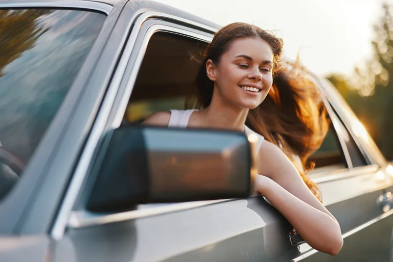 Happy young woman enjoying a road trip with her hair blowing in the wind from a car window during sunset, wearing a casual outfit and smiling brightly.