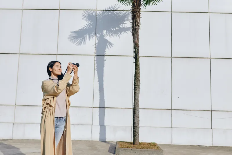 Woman photographs palm tree against a white tiled wall in bright outdoor light, camera raised, stylish coat and jeans, urban setting, candid lifestyle scene and photography action
