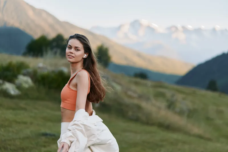 A relaxed woman stands in a grassy meadow with rolling hills and distant mountains, wearing an orange top and light jacket, smiling softly as sunlight warms the landscape.