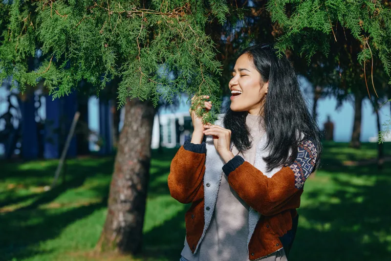 Woman smiling outdoors, reaching for tree branches in a sunny park, casual sweater and joyful mood captured among greenery, a candid nature scene perfect for lifestyle and outdoor photography