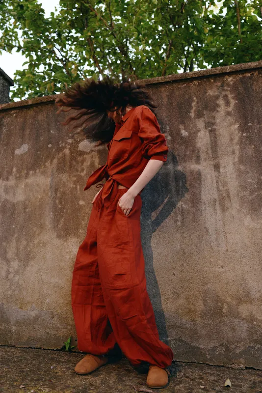 A woman in a burnt orange jumpsuit poses outdoors beside a weathered wall, hair flipping with movement, showcasing confident street fashion and a relaxed lifestyle, natural light highlighting texture