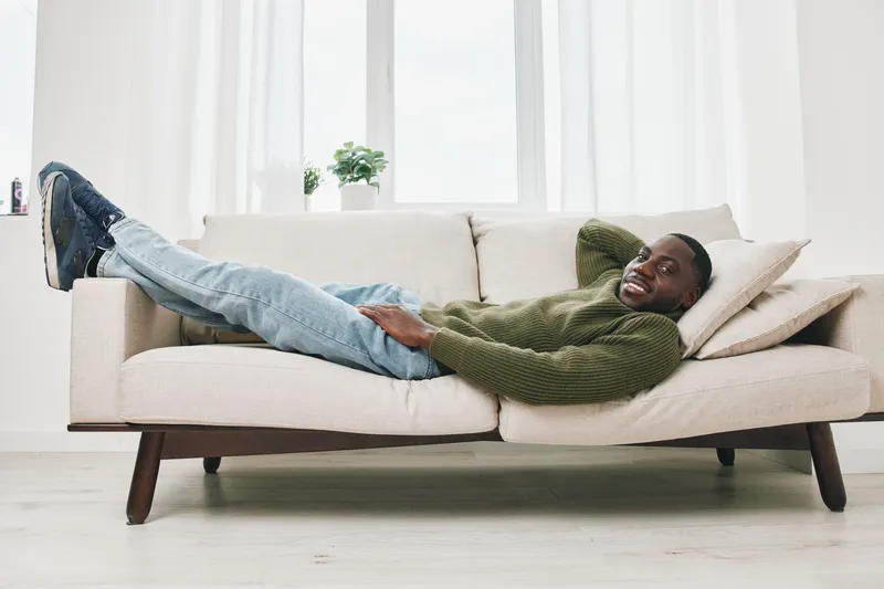 Relaxed young man lounging on a modern couch, wearing casual attire, smiling with a comfortable expression in a bright room with a minimalist design