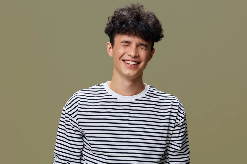 smiling young man in striped shirt with curly hair laughing joyfully against simple olive background, casual happiness, positive expression, ethnic diversity