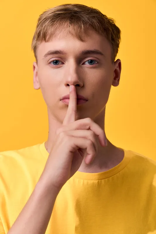 Shushing, silence, discreet secret gesture by a youth man with finger on lips, yellow background, studio setting, calm mood, quiet communication and minimal style for secrecy