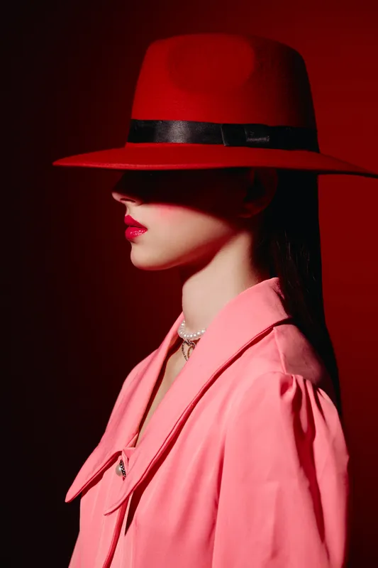 Elegant woman in a vibrant red hat and soft pink coat poses in profile against dark background. Modern fashion look highlights makeup and pearl necklace in creative beauty portrait.