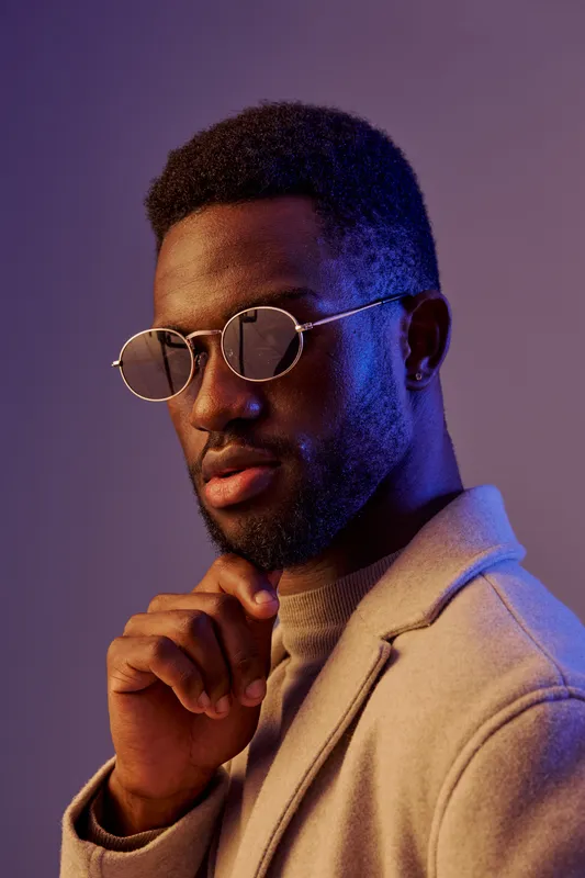 A stylish young man wearing round sunglasses and a beige coat, exuding confidence with a thoughtful expression against a colorful, gradient backdrop