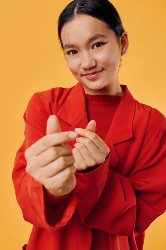 Young woman in red jacket shows heart gesture with hands smiling at camera on orange background for love, emotion and gesture expression in studio portrait.