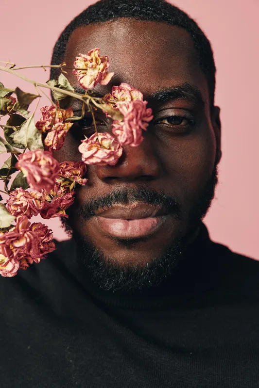 Creative conceptual portrait of African man with dried pink flowers covering one eye, wearing black turtleneck against soft pink background in visual artistic style.