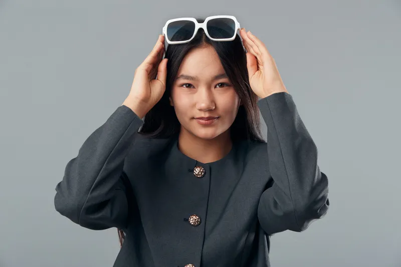 Young woman with long black hair wearing dark jacket adjusting white sunglasses on head against gray background. Fashion portrait with confident expression and modern style casual clothing.