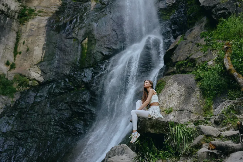 Woman sitting near waterfall in white activewear with relaxed and peaceful expression surrounded by lush green vegetation and rocky terrain outdoors.