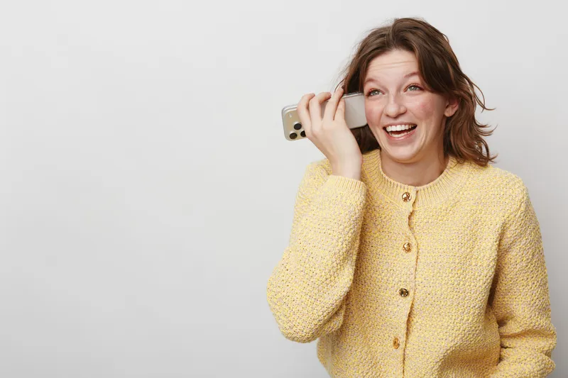 Happy young woman in yellow sweater talking on smartphone, smiling and looking away. Isolated on light background. Concept of communication, technology, and positive emotions.