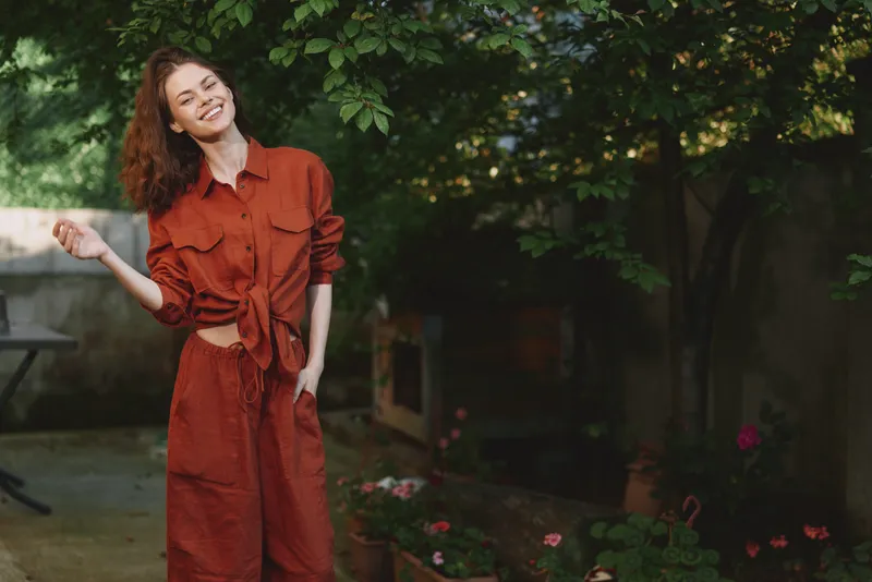 Woman lifestyle portrait outdoors wearing a red jumpsuit, smiling with playful pose in a sunny garden, conveying relaxed confidence and cheerful mood, candid fashion moment for photographers