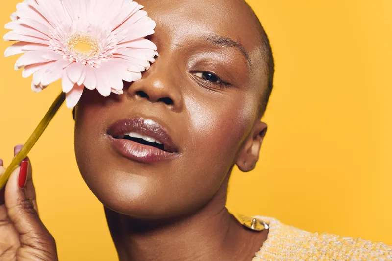 Close-up of a smiling black woman with dark skin holding a pink flower near her face against a yellow background. Concept of natural beauty, skincare, happiness, and wellness.