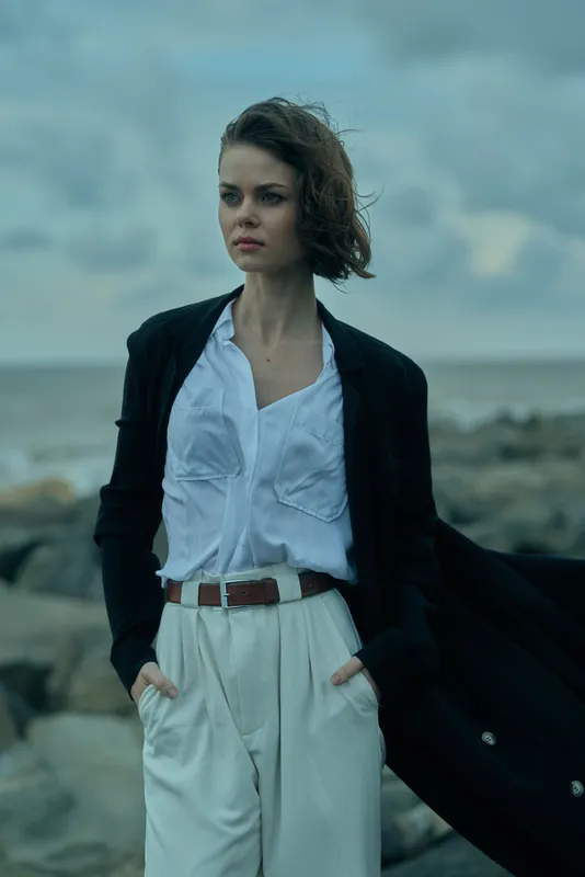 Woman on a windy coast wearing a white shirt, light trousers and a dark coat billowing in the breeze, moody fashion scene by the sea, elegant coastal style.