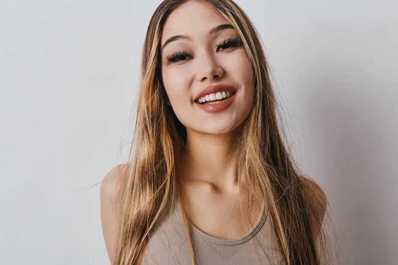 Young woman with long hair smiling brightly with teeth visible, wearing sleeveless top and looking at camera against plain light background for portrait photography.