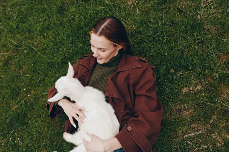 A woman in a brown coat sits on grass, cradling a white goat kid with a gentle smile. An outdoor lifestyle moment that captures calm bonding with animals.
