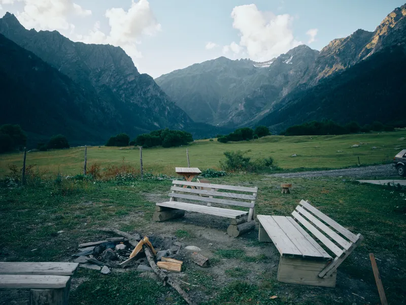 Mountain valley scene with weathered wooden benches around a campfire pit, green meadow and distant peaks under a clear sky, inviting contemplation and outdoor escape