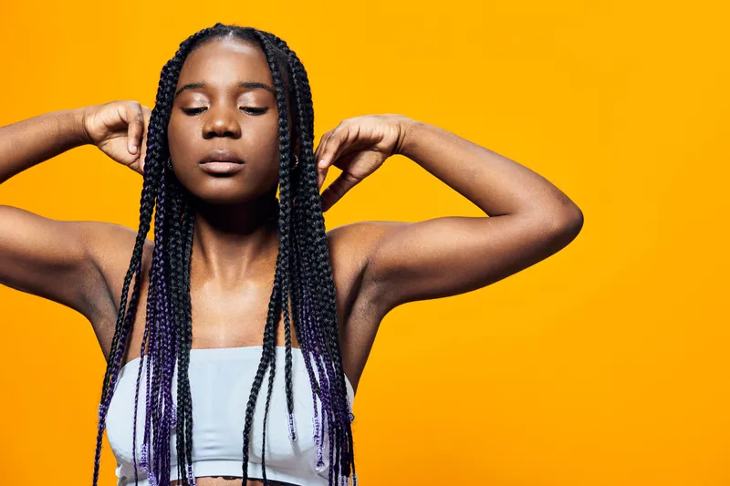 Young Black woman with braids relaxing against a vibrant yellow background, expressing tranquility and confidence through her body language and joyful demeanor
