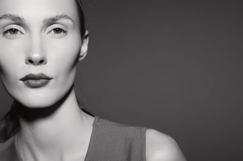 Close-up of a pale white European woman with slicked hair and neutral expression against dark grey background in monochrome studio portrait.