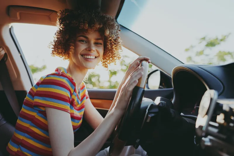 Bright outdoor moment with a smiling woman behind the wheel in a striped shirt, casual driving scene, sunlit interior, cheerful vibe and natural colors, warm breeze, candid pose, vibrant film color