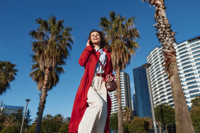 Young woman in red coat walking outdoors on sunny day, smiling happily, palm trees and modern buildings in background, casual outfit, urban lifestyle scene
