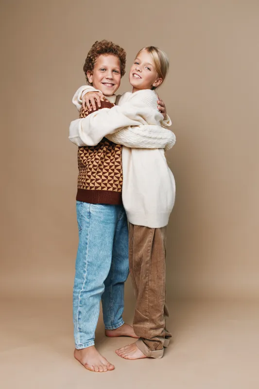 Two children embrace warmly, smiling, wearing cozy knitwear and jeans against a soft tan backdrop, capturing friendship, comfort, and playful innocence.