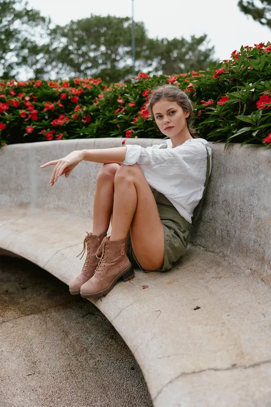 Young woman in stylish outfit with shorts and boots sitting on a bench, surrounded by vibrant flowers, exuding confidence and casual elegance