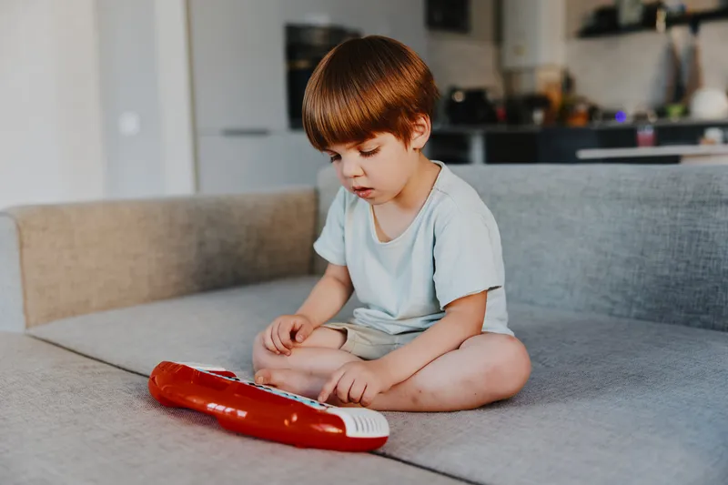 Curious boy playing with a toy keyboard, sitting cross-legged on a sofa. The child is focused, exploring music in a cozy living room setting.