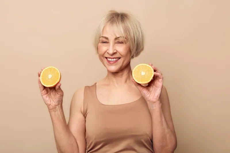 Happy mature woman with short blonde hair holding two halves of fresh orange fruit, smiling with eyes closed over beige background. Concept of healthy lifestyle, vitamin C, and wellness.