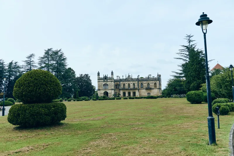 Castle in a manicured park sits beyond a green lawn and shaped trees, with a classical lamp post along a curved path, creating a serene historic landscape scene