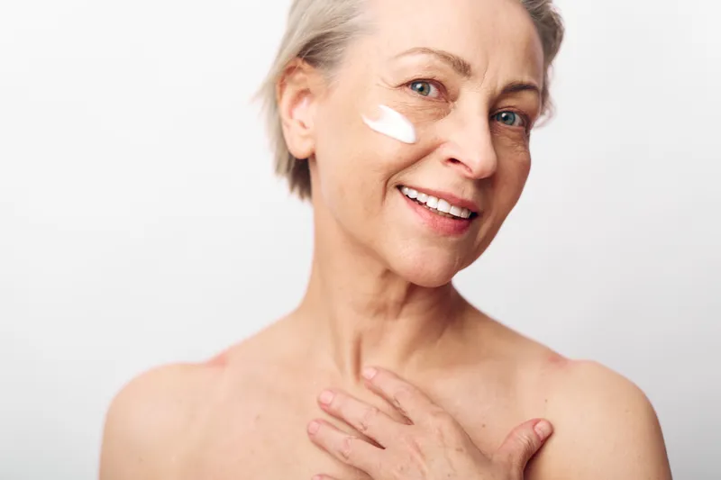 Mature woman with short hair applies white face cream on cheek. Close-up portrait on white background. Concept of skincare, aging skin care, beauty routine, and self-care.