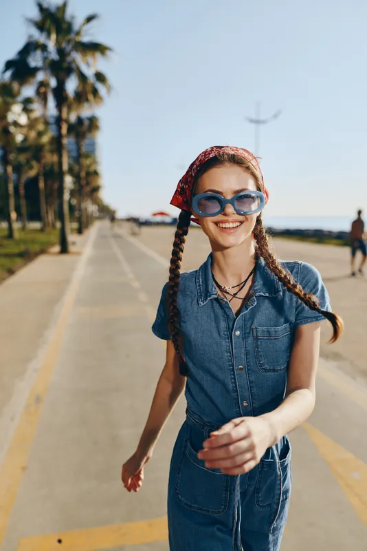 Lifestyle woman in modern boho-western denim street style smiling joyfully outdoors, wearing sunglasses and a red bandana with bright film color tones.