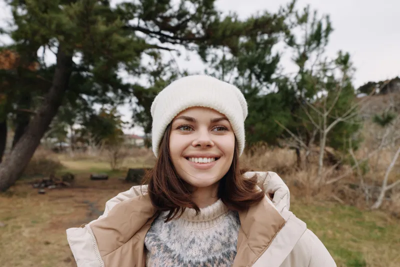 Woman outdoor smiling portrait in a winter jacket and beanie, enjoying a chilly day in a park with trees and open space for lifestyle photography.