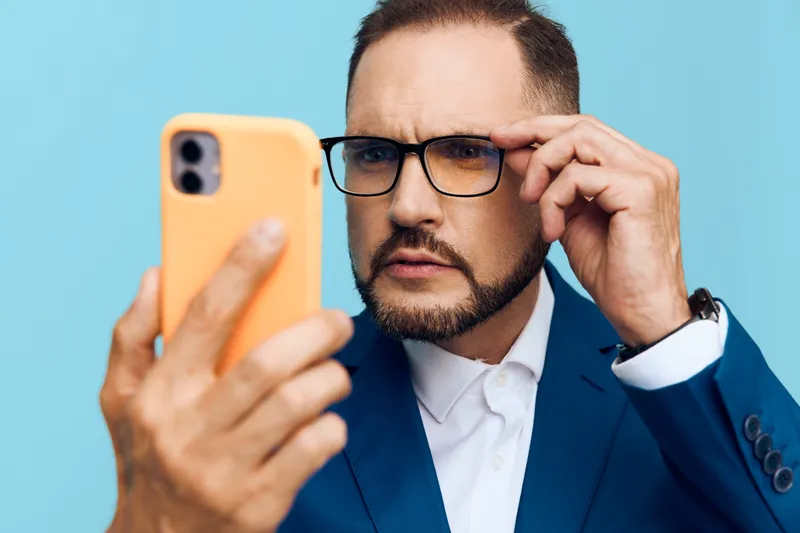 A professional man in a blue suit on a solid colored background, looking at a smartphone with a focused, confident expression.