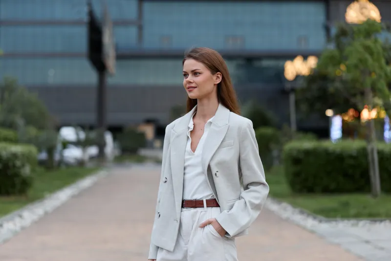 Confident businesswoman in a stylish light blazer and pants walking in an urban setting with a soft smile, showcasing professionalism and elegance in her appearance.