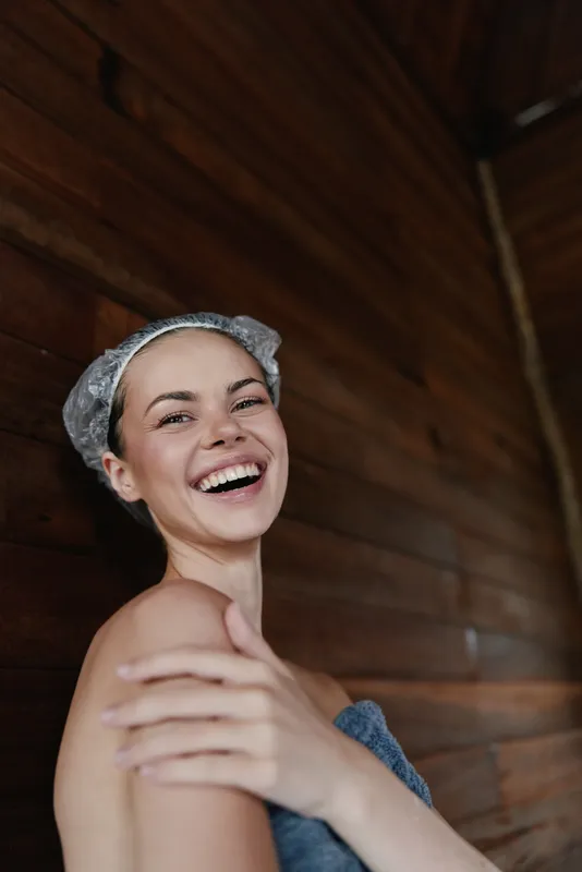smiling woman wrapped in towel with shower cap expressing happiness and freshness against wooden background indoors, natural skincare and bath concept