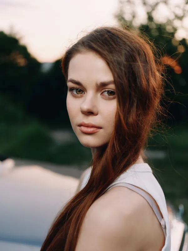 Young woman with long brown hair gazing thoughtfully, dressed in a light summer outfit, against a vibrant sunset background, conveying a sense of serenity and reflection.
